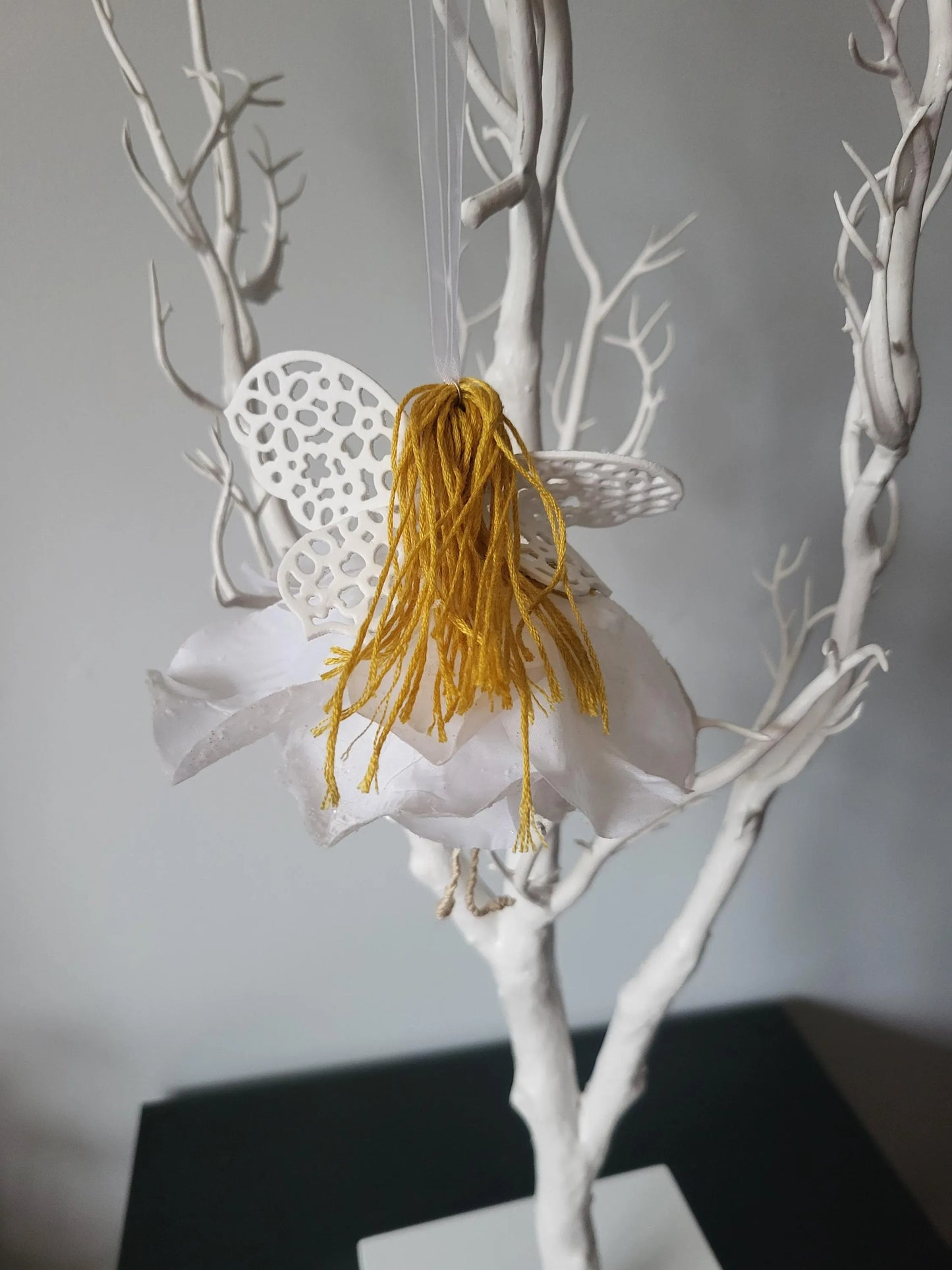 Artistic representation of a fairy with golden hair and white lace wings on a branch against a grey background