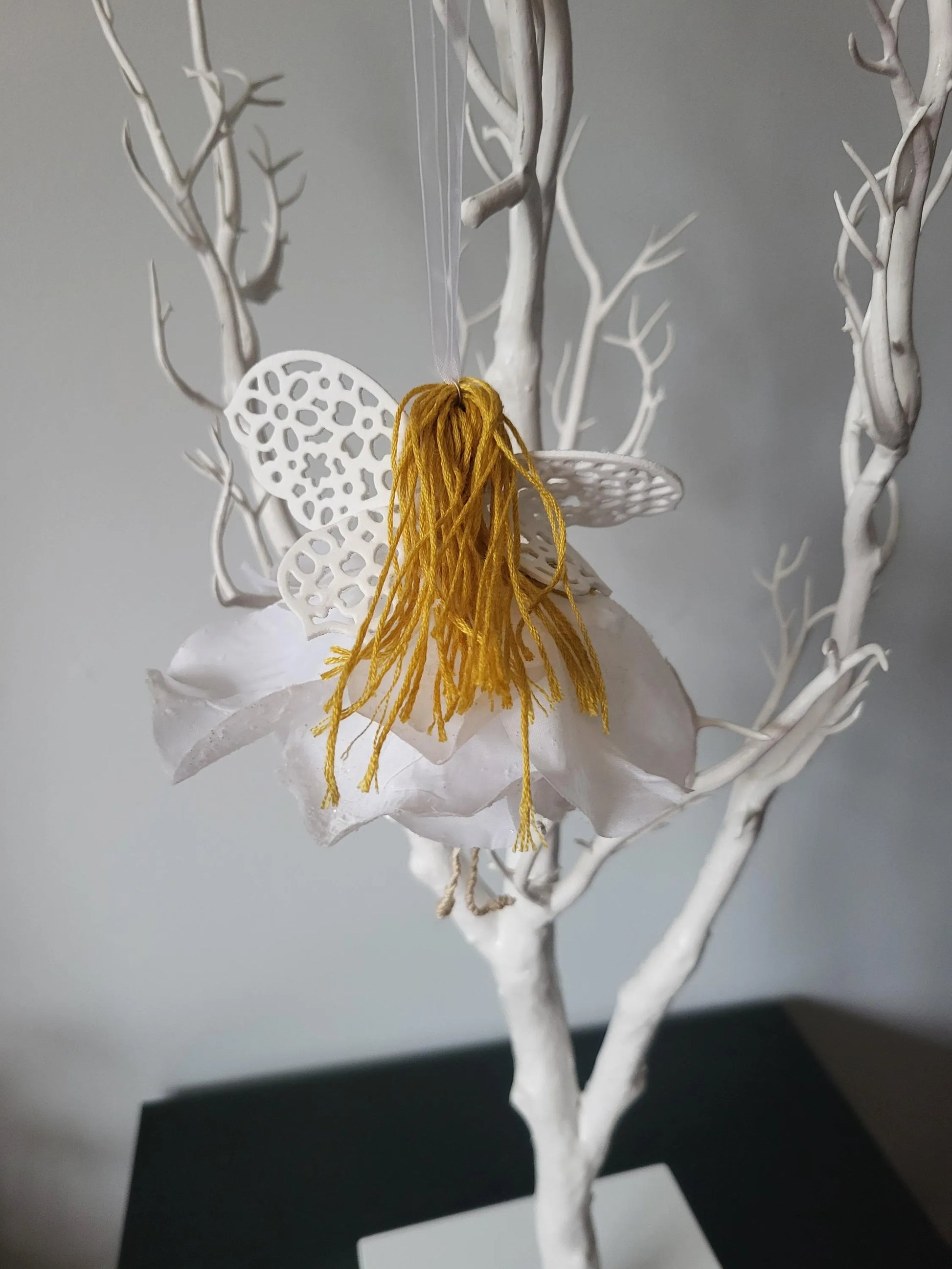 Artistic representation of a fairy with golden hair and white lace wings on a branch against a grey background